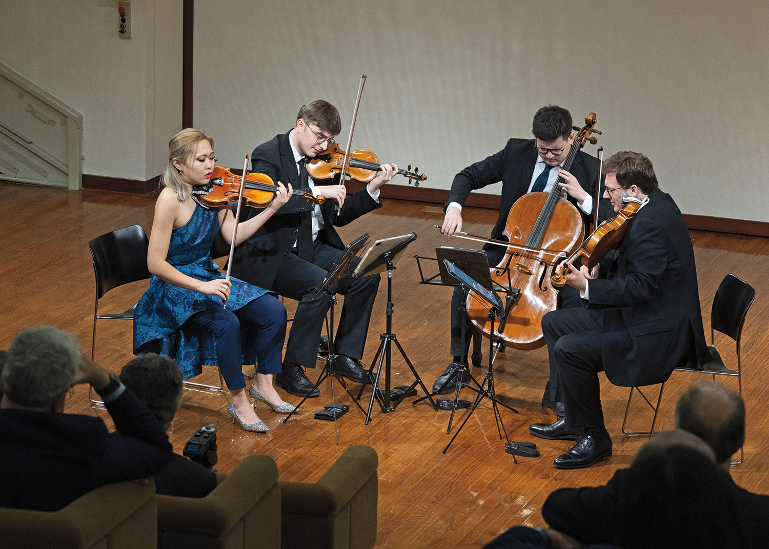 Four musicians sit in a semi-circle onstage facing music stands and sheet music. They are dressed in formal attire and play violas, violons, and violoncellos for a small audience.