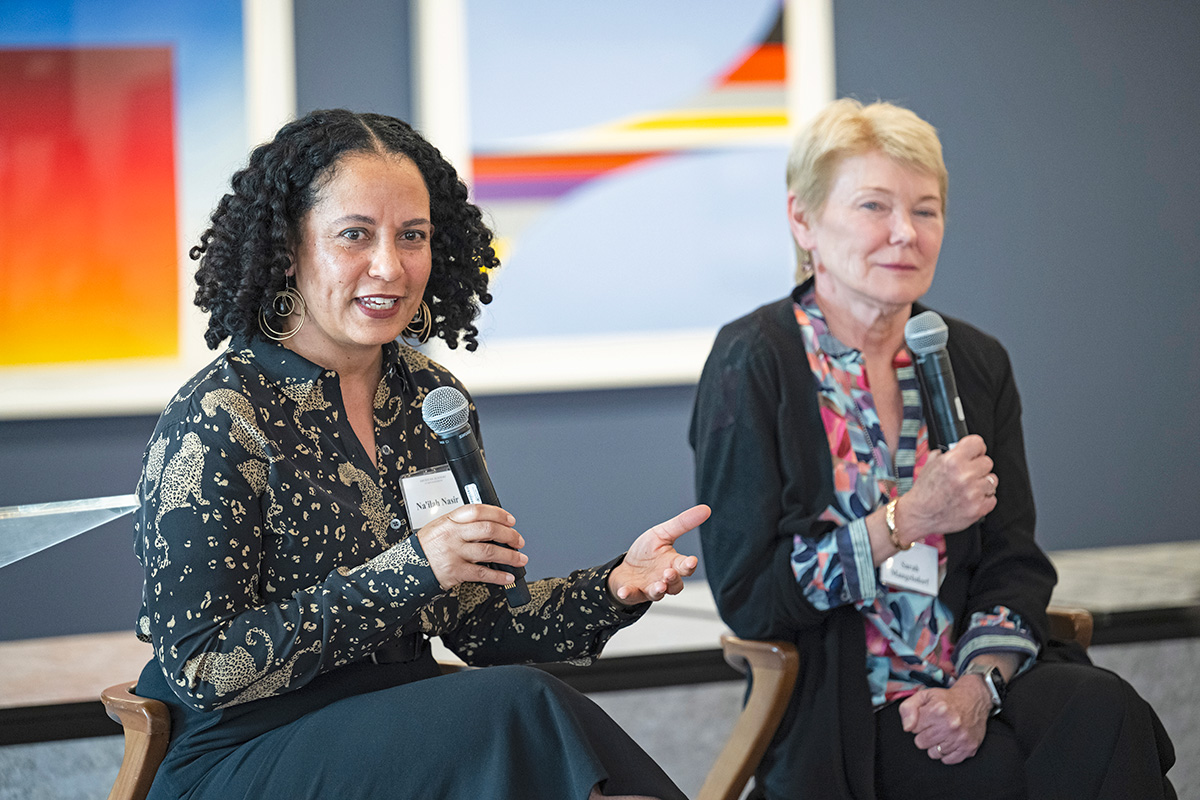 Two women are sitting on stage, holding microphones. The woman on the left, with brown curly hair and brown skin, is speaking. The woman on the right, with light skin and short blond hair, is listening.