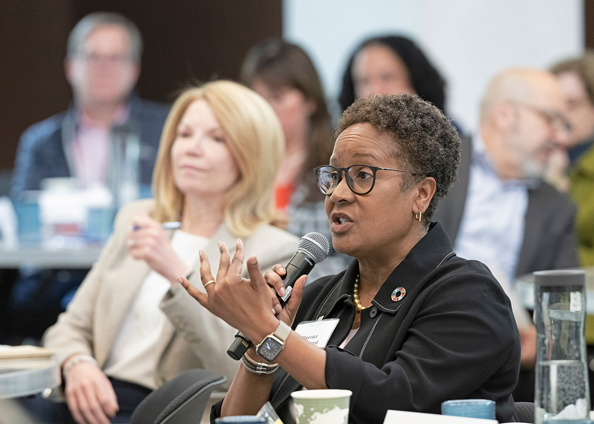 A woman, with short black curly hair and dark skin, is seated at a table, holding a microphone in her right hand and speaking into the microphone. To her right is a woman with light skin and long blond hair, who is seated and listening.