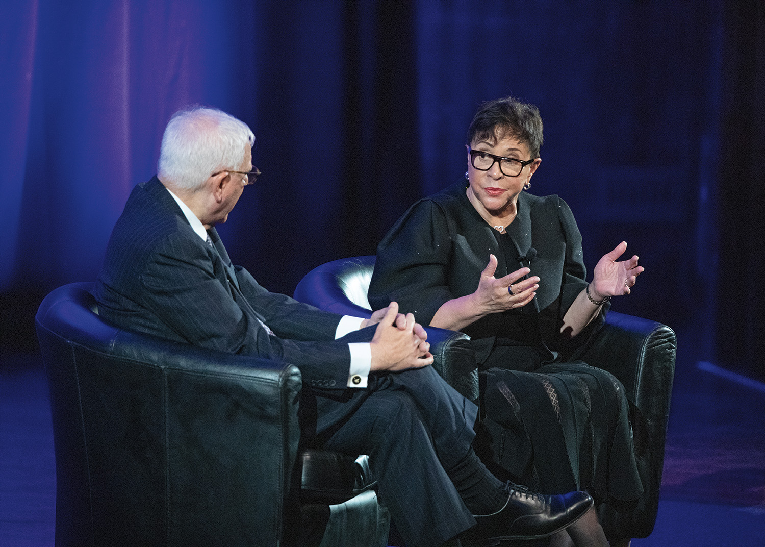 David Rubenstein and Sheila Johnson sit on a stage in large black chairs during an event at the 2023 induction ceremony. David Rubenstein wears a striped suit and glasses and has short, white hair, and pale skin. Sheila Johnson wears a black dress, coat, and glasses, and has short, black hair and light brown skin.