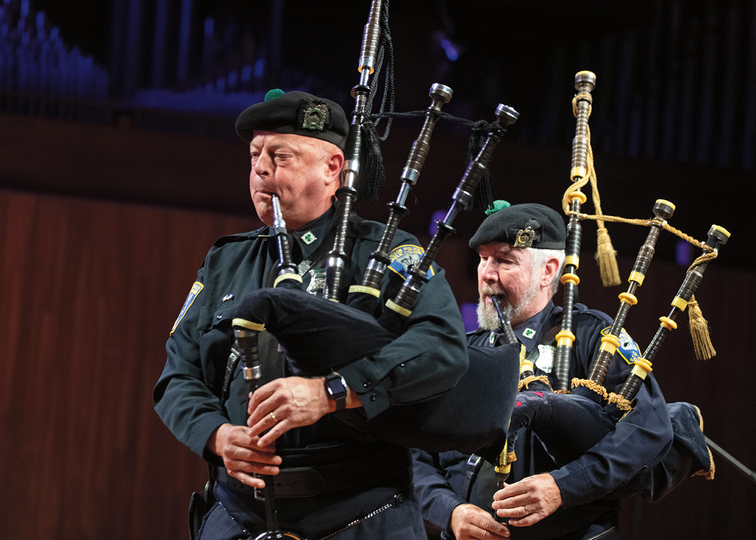 On the left, a person with pale skin plays a bagpipe. His cheeks are puffed. He wears a police uniform with badges and pins. On his head is a black beret with a green pom-pom on top. On the right, a person with pale skin and a white beard plays a bagpipe. He bites one of the reed pipes. He wears a police uniform with badges and pins. On his head is a black beret with a green pom-pom on top.