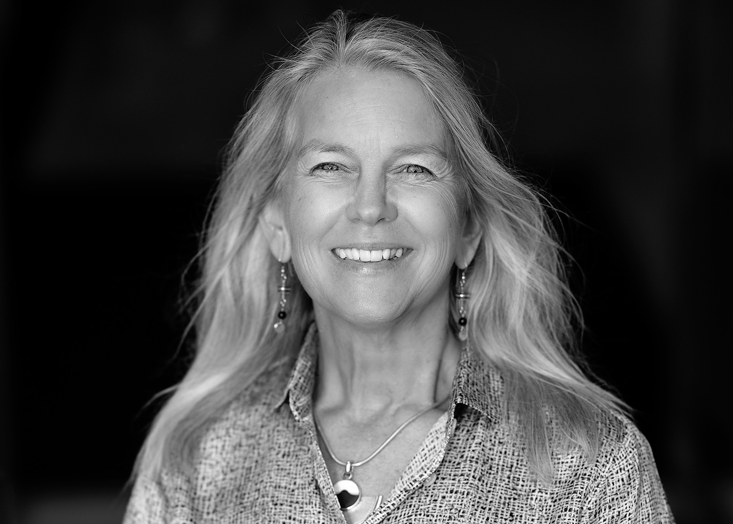 A black and white headshot of Dava Newman. She faces the camera with a big smile. She has long hair. She wears hanging earrings, a necklace with circular shapes, and a patterned button-down shirt.