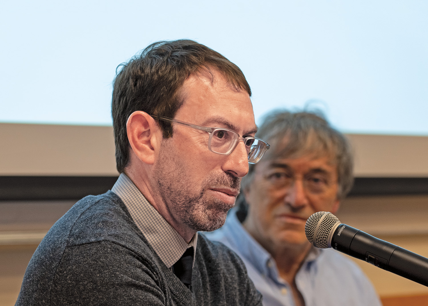 Samuel Moyn sits in front of a microphone. He wears a sweater, collared shirt, and glasses. Moeshe Halbertal sits in the background and watches him talk.