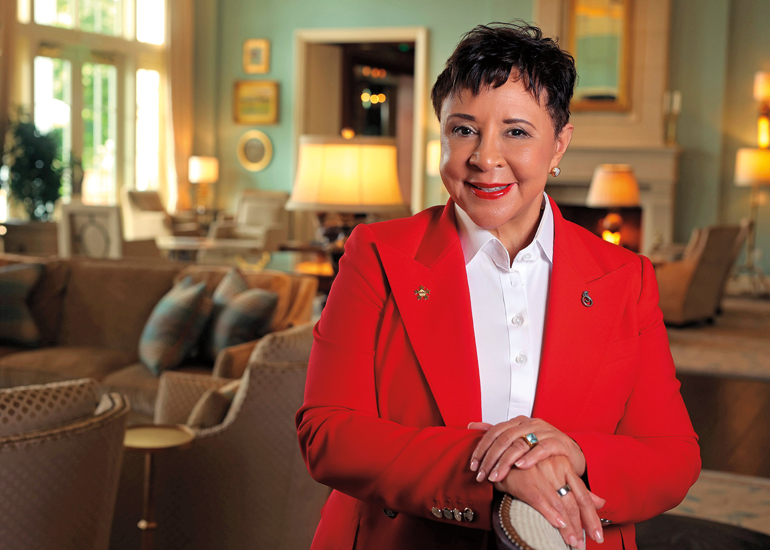 A headshot of Sheila Johnson. She faces the camera and smiles. She has light brown skin and short black hair. She wears earrings with diamonds and gold, a white collared shirt, and a red suit jacket with pins on the lapels.