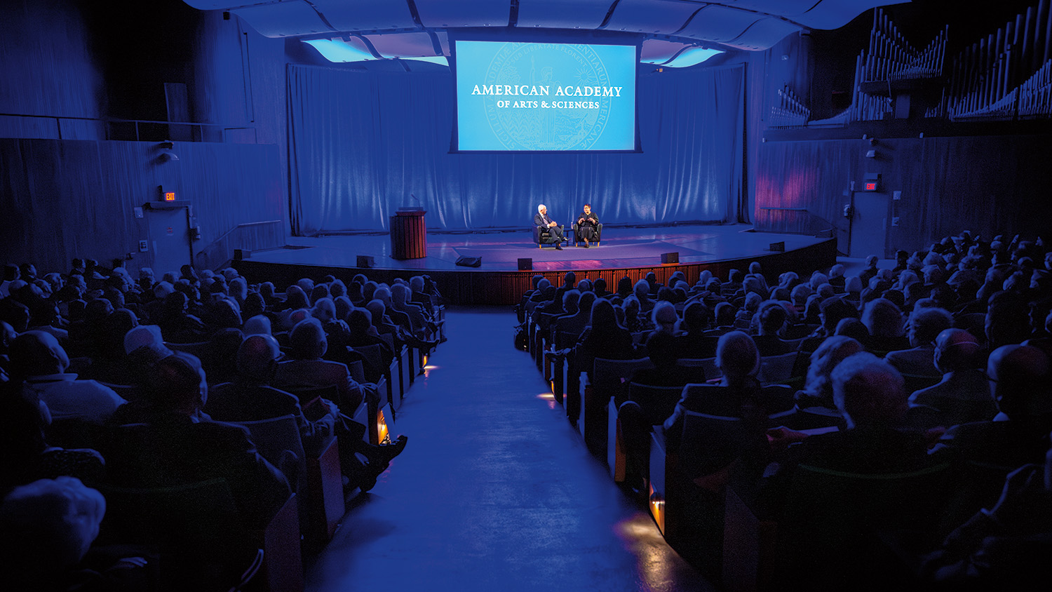 A large audience watches two people talk on a stage. The audience is in the dark. The people sit in large chairs on the stage. They are bathed in blue lights and surrounded by blue curtains. Behind them, a projection screen reads American Academy of Arts and Sciences.