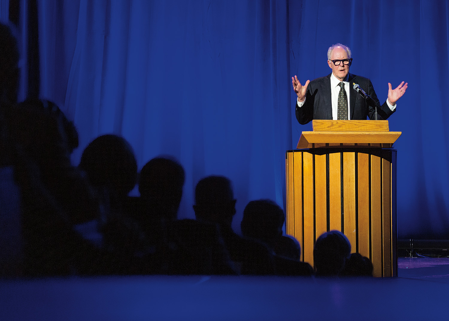 Actor John Lithgow stands onstage at a podium at the 2023 induction ceremony. He faces the audience with his hands raised. He has pale skin and grey hair. He wears glasses and a dark suit with a white shirt and tie.