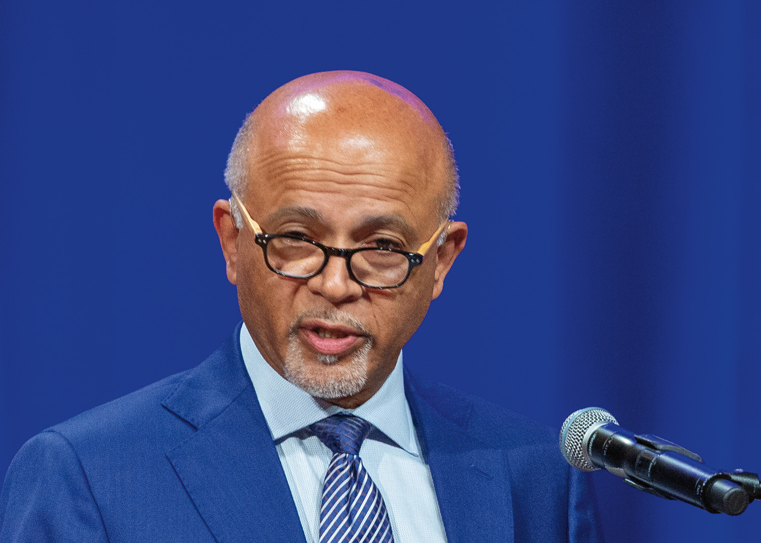 Abraham Verghese stands at a podium in front of a microphone. He has short, white hair and light brown skin. He wears glasses, a light blue collared shirt, a tie with red, white, and blue stripes, and a dark blue suit.
