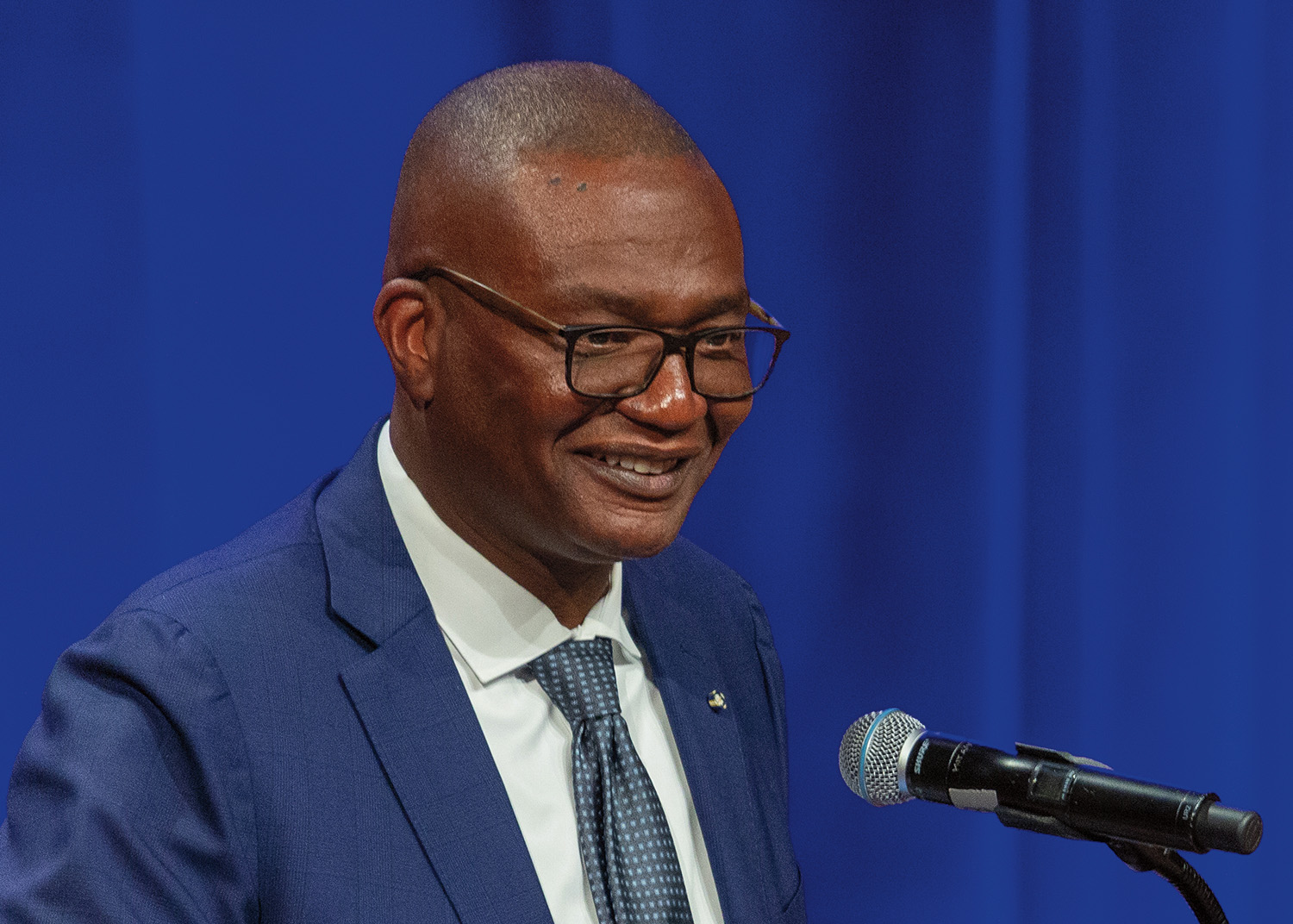 Kerwin Charles stands at a podium in front of a microphone. He smiles. He has short, black hair and dark brown skin. He wears glasses, a white collared shirt, a tie with tiny blue squares, and a dark blue suit with pins on the lapel.