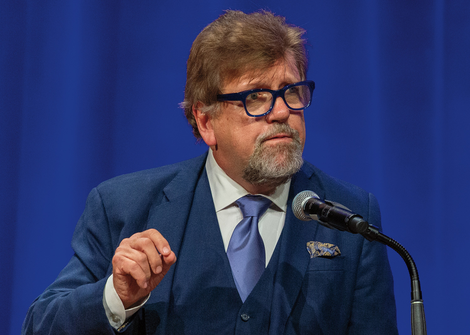 Oskar Eustis stands at a podium in front of a microphone. He has short, wispy brown hair and pale skin. He wears blue glasses, a white collared shirt, a light blue tie, and a dark blue three-piece suit.