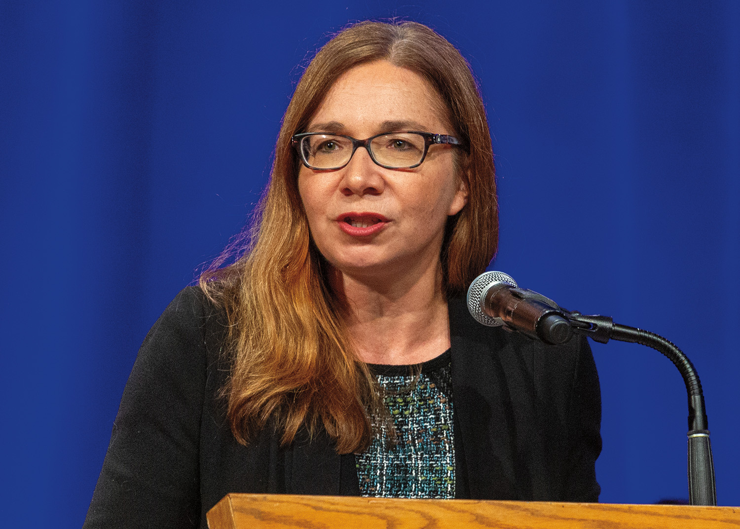 Katharine Hayhoe stands at a podium in front of a microphone. She has long, brown hair and pale amber skin. She wears a dark tweed dress and black cardigan.