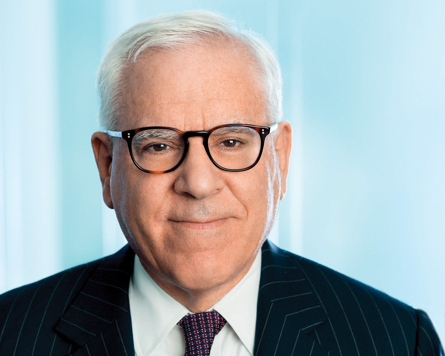 A headshot of David M. Rubenstein. He faces the camera and smiles. He has pale skin and short white hair. He wears plastic glasses, a blue and red tie, a white collared shirt, and a blue pinstriped suit. Photo courtesy of David M. Rubenstein.