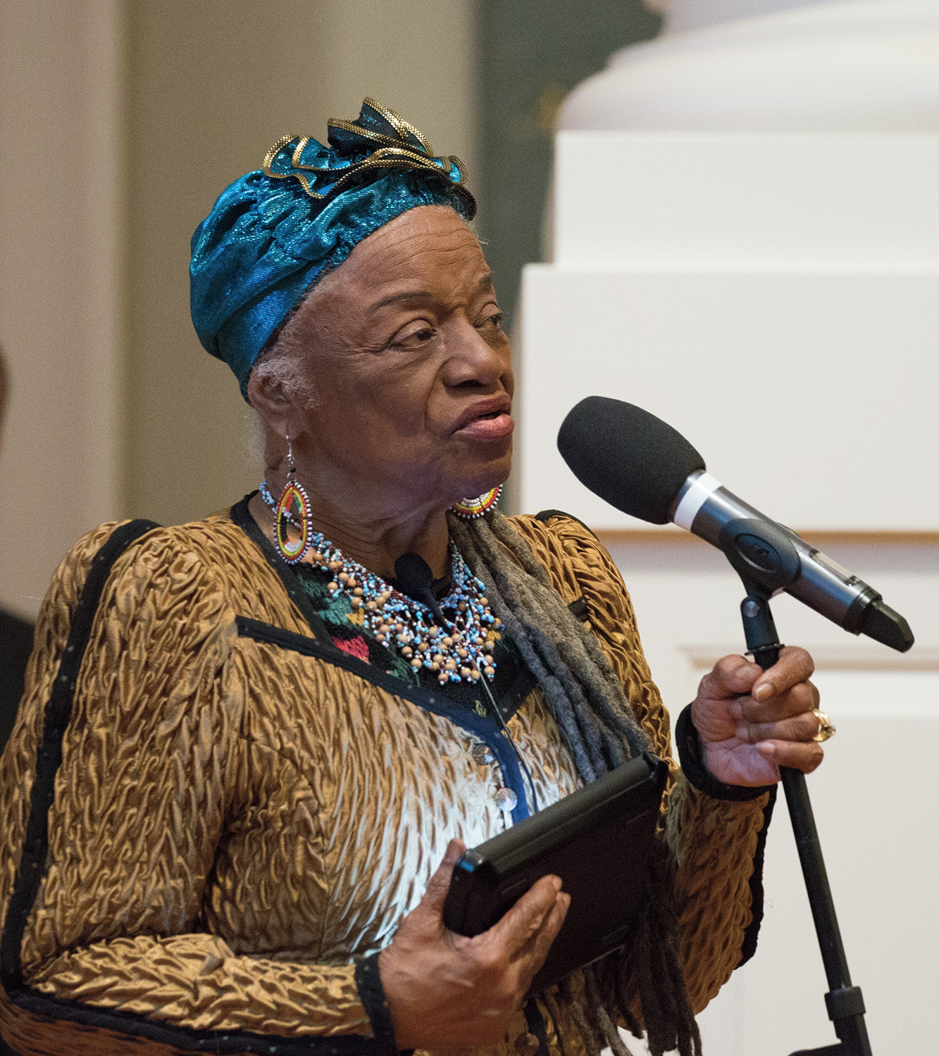 A photograph of Faith Ringgold, a dark-skinned elderly person wearing a blue head wrap, layered necklaces in blue and beige, and a beige festive blouse. Ringgold is speaking into a microphone while holding a computer tablet.