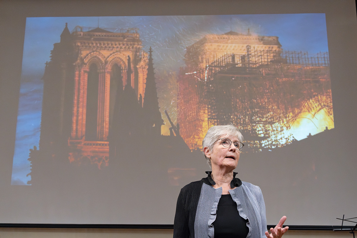 A woman presents in front of an image of Notre-Dame Cathedral burning. 