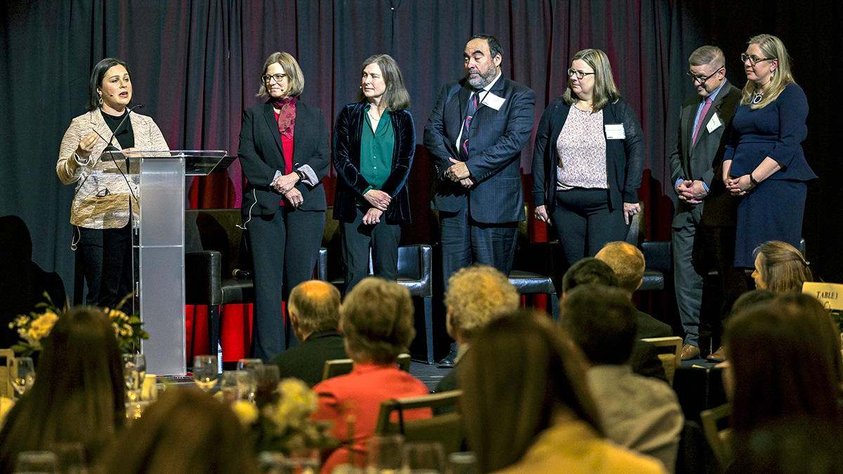 A group of six people stands on a stage facing an audience, with one person speaking at a podium.