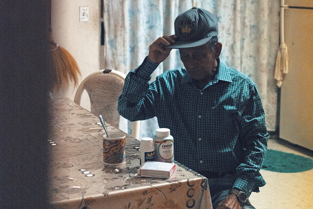 A person with short grey hair and brown skin seating at a kitchen table looking at medicine. Their head is down and their face is covered by the shadow of their hat. 