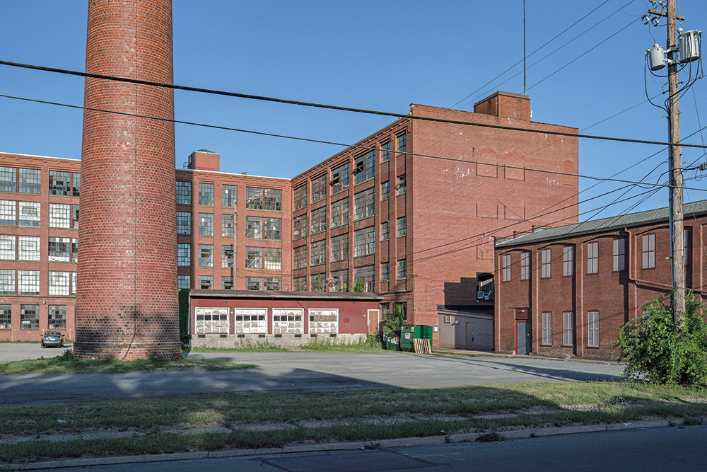 A red brick building and smokestack of an old factory with a blue sky in the background. Some building windows are open or broken. One car sits in the parking lot. 