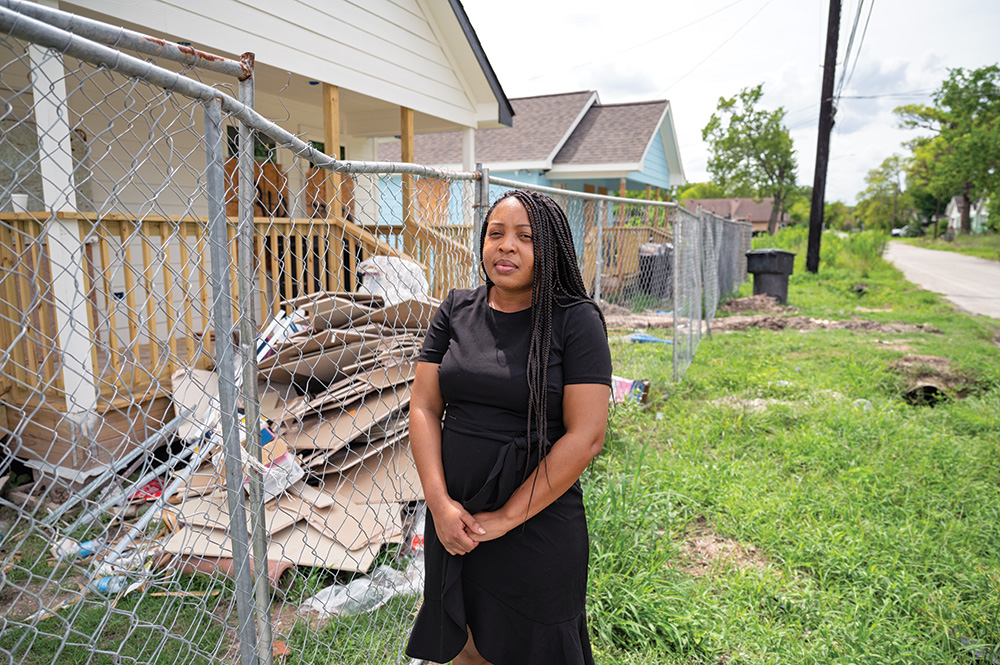 A person with brown skin with braids stands with their hands folded in front of them. They are wearing a black dress and look toward the viewer. They stand on a stretch of grass between a chainlink fence and a road. Behind the fence, old houses are boarded up and various building materials piled across their lawns.