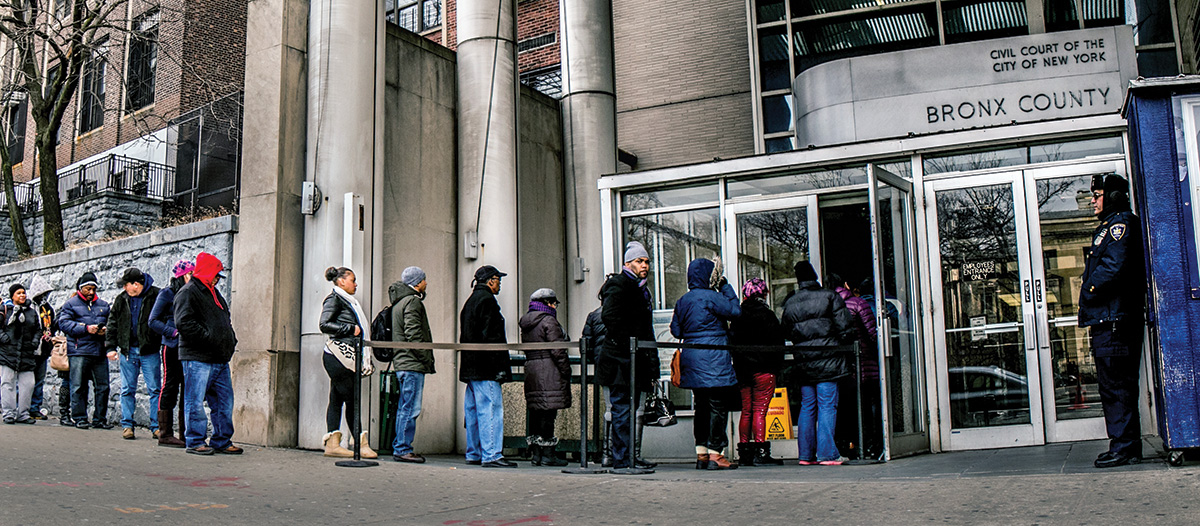 A line forms outside of the Bronx county housing court. People stand outside the court in jackets as a security guard watches the courthouse entrance.