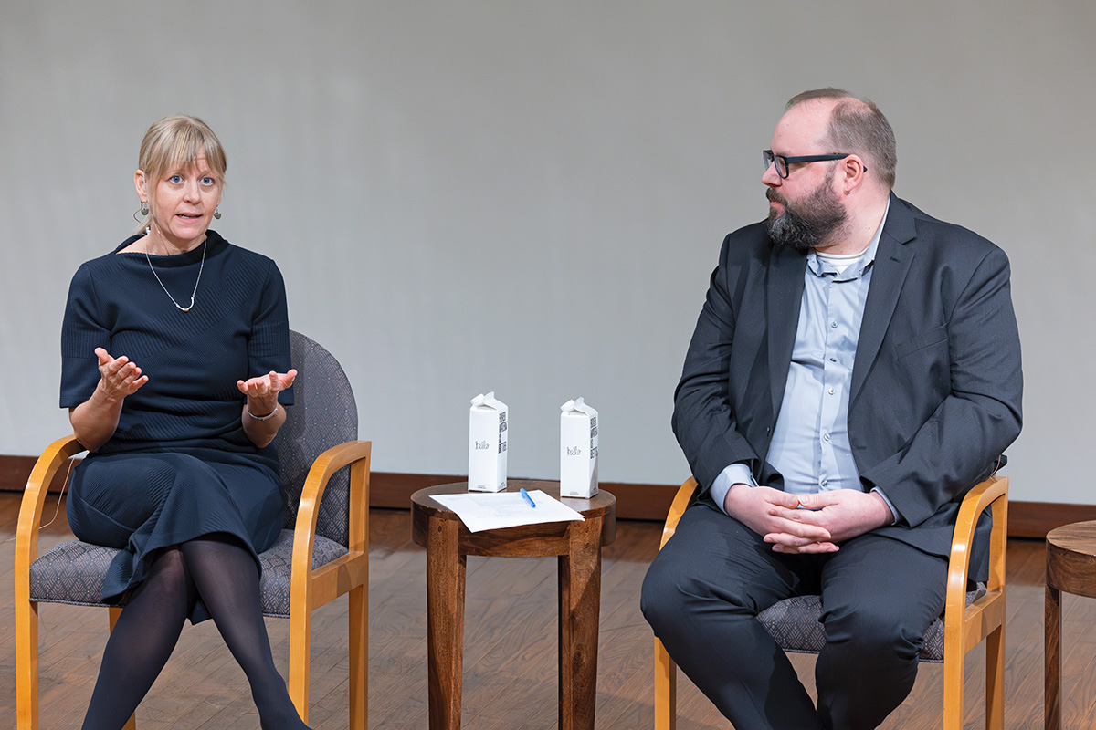 Nikole Nelson sits beside Matthew Burnett, facing an audience (off-camera), while Matthew turns to face her as she speaks. A paper, pen, and two boxes of water are on a small wooden table between them. Both Nikole and Matthew wear business attire. Nikole has pale skin and long blond hair. Matthew has pale skin and short dark hair.