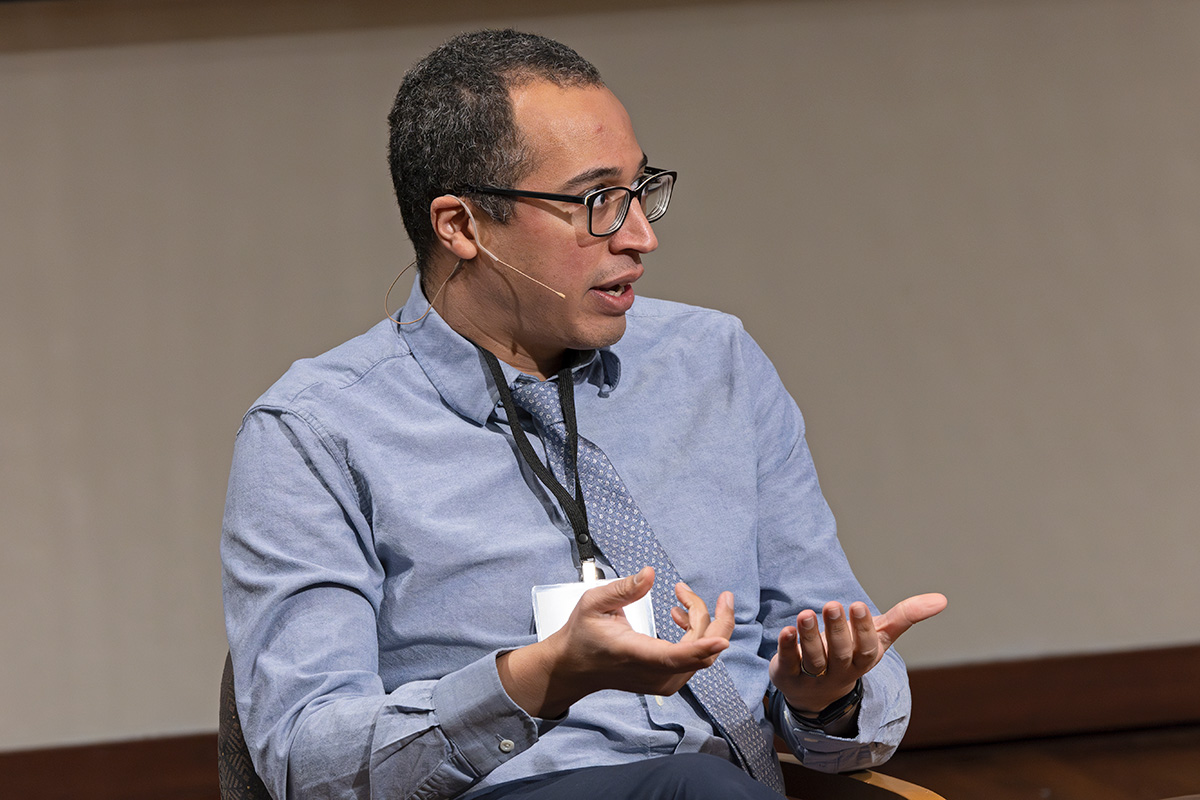 Quentin Steenhuis wears a headset microphone and speaks to someone off-camera. Quentin has brown skin and brown hair, and wears glasses and business attire.