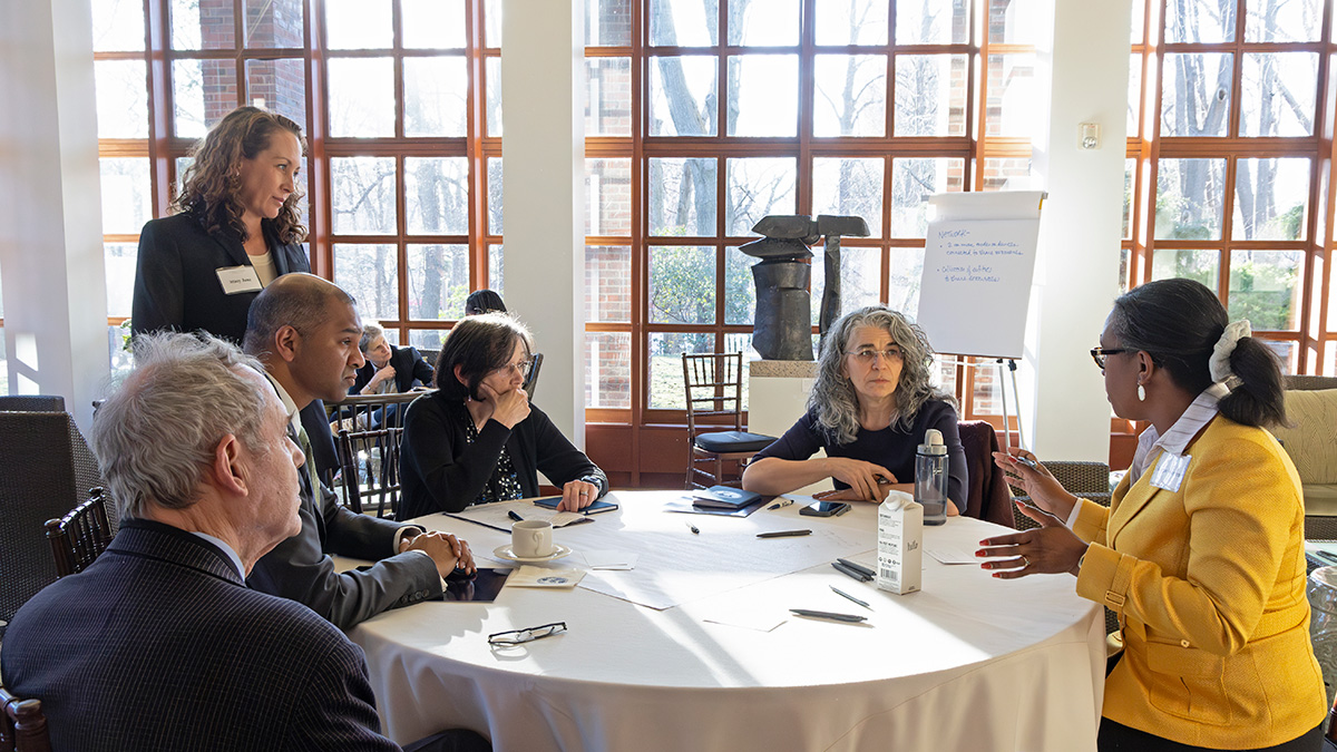 A facilitator stands and observes a group seated around a table, engaged in conversation at the Making Justice Accessible Summit. The facilitator has pale skin and long auburn hair. The group includes people of various races and genders.