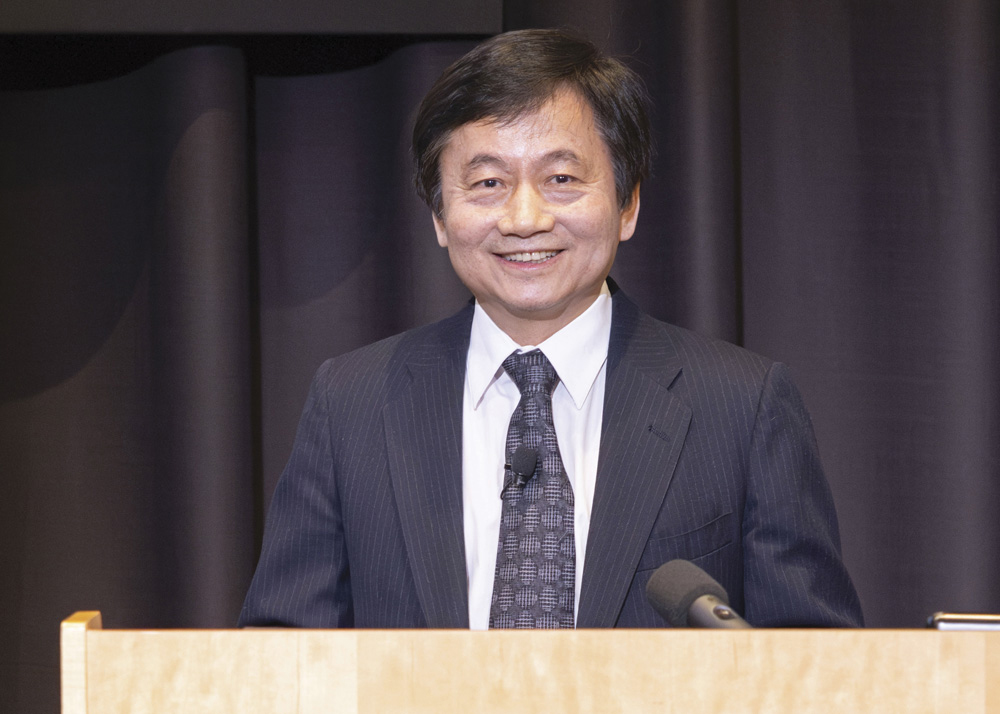 A man with dark hair, wearing a dark suit jacket, white shirt, and dark tie, stands at a podium and smiles at the viewer. 