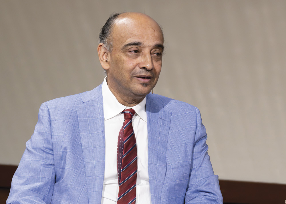 A man with dark skin, wearing a light blue suit jacket, white business shirt, and red tie with blue stripes, is in conversation with a person to his right (not shown).