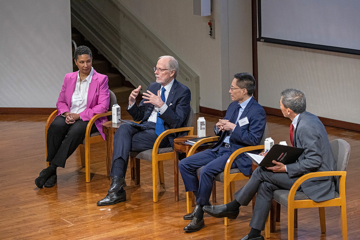 Four individuals are seated next to each other and facing an audience that is not shown.  The second person from the left is speaking and has his hands raised to emphasis a point he is making.  The other three individuals are turned and facing the person who is speaking.