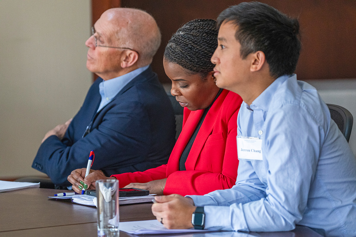 A man with pale skin in a blue jacket, a woman with dark skin in a red jacket, and a man in an Oxford blue shirt take notes and listen to a speaker who is off camera.