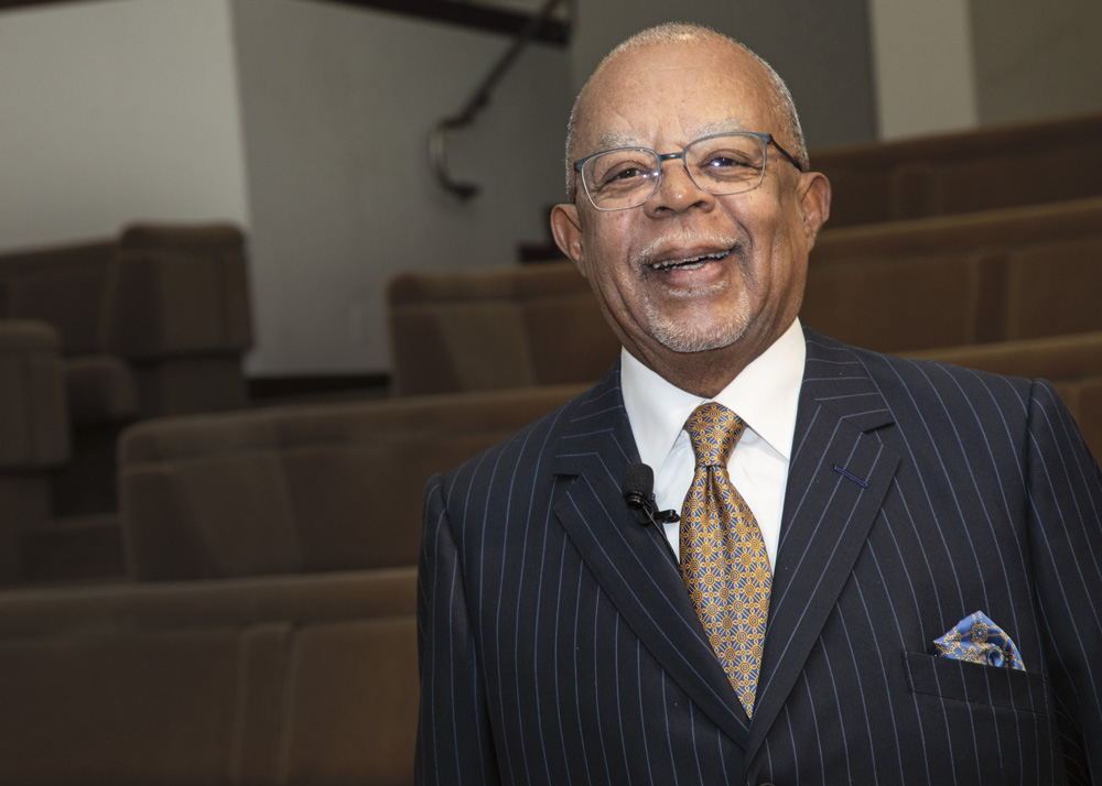 A man with dark skin, wearing a dark blue pinstriped jacket, white business shirt, and yellow and blue paisley tie, smiles at the viewer.