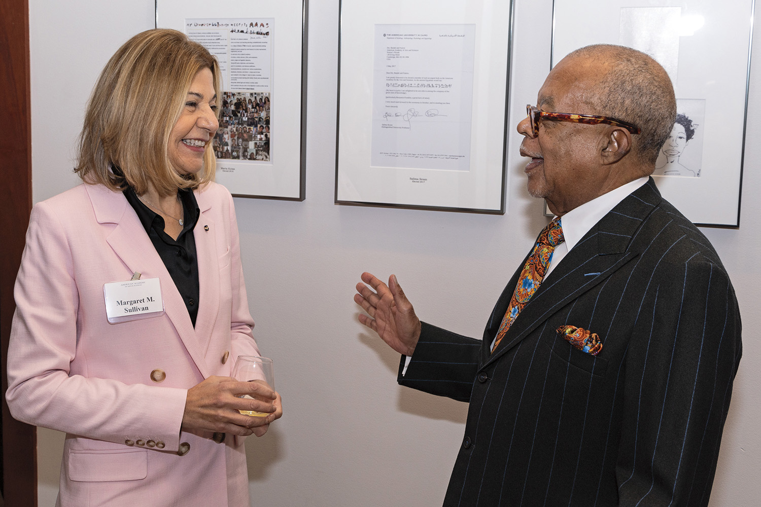 Two people wearing business suits have an animated conversation in front of framed letters written by American Academy members. 