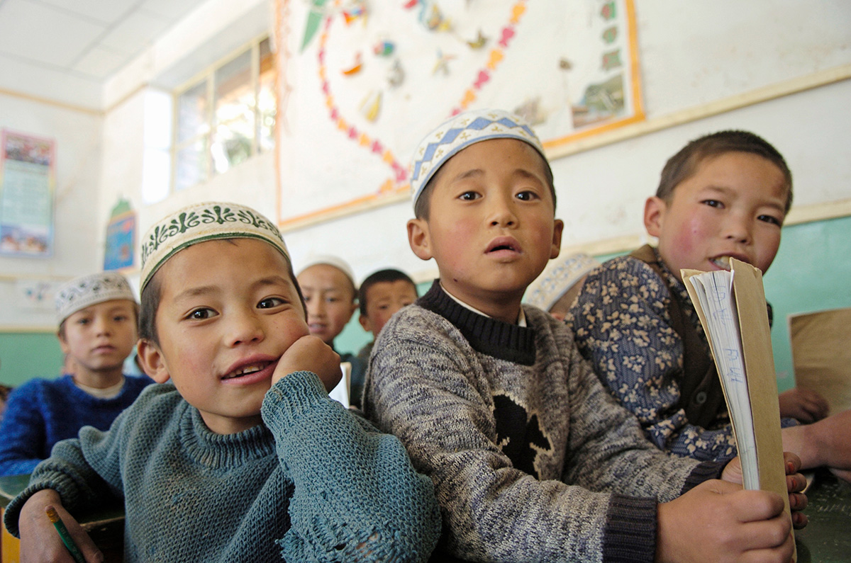 Six young students sit in a classroom in Gansu province, China. They look toward the front of the class. One student holds a book. The students have light brown skin and black hair. Many wear short white hats traditional among Salar people.