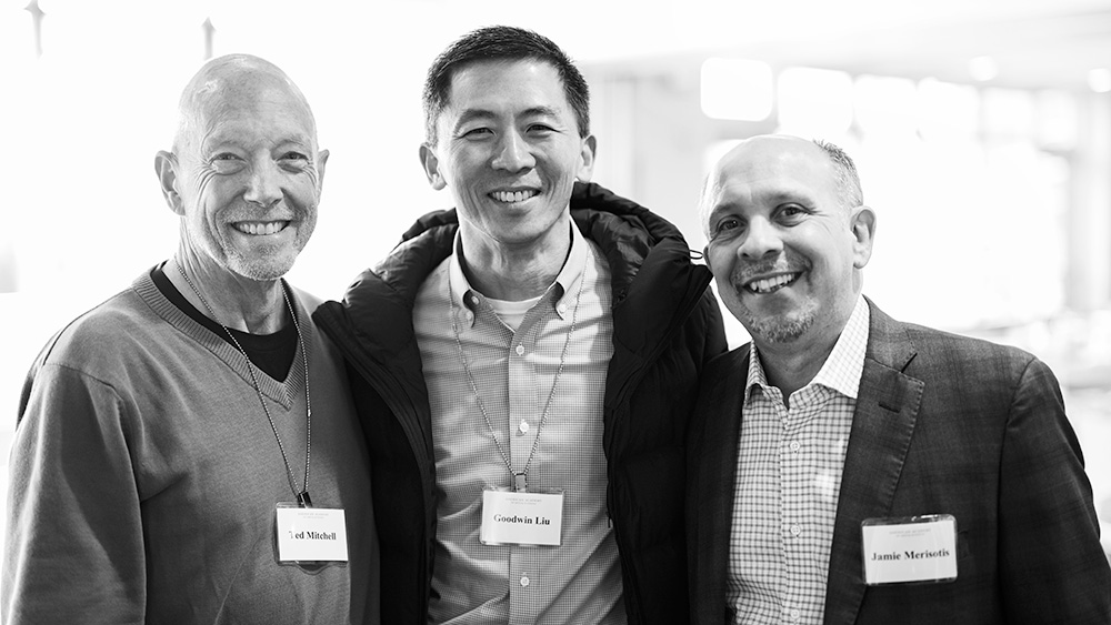 A black and white portrait of Ted Mitchell, Goodwin Liu, and Jamie Merisotis facing the camera and smiling. Mitchell has pale skin and is bald. He wears a sweater over a dark shirt. Liu has short black hair and light skin. He wears a dark coat over a checkered shirt. Merisotis has pale skin and short graying hair. He wears a gray suit and glasses.
