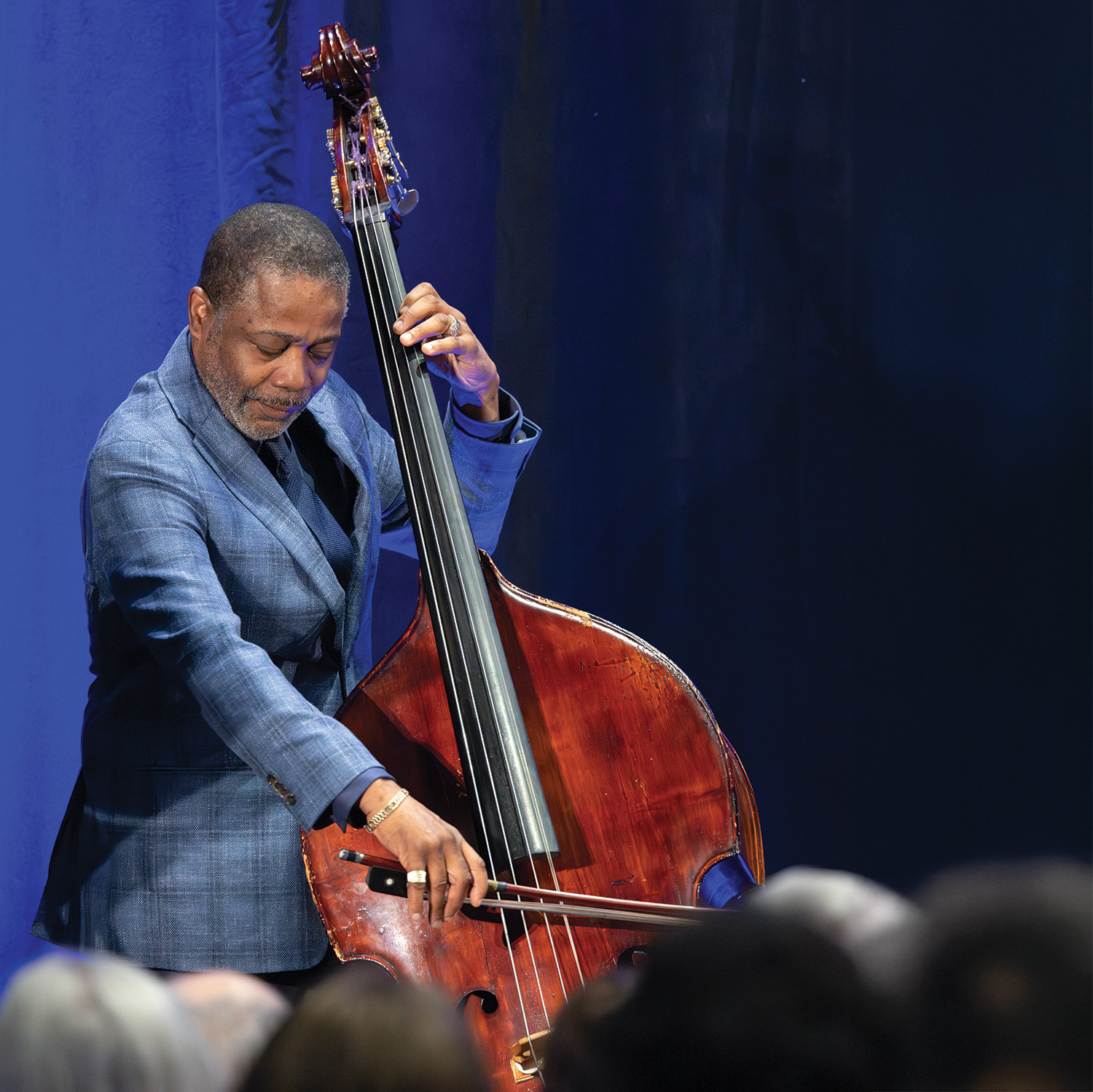 A dark-skinned man, wearing a blue suit jacket and tie, plays the double bass.