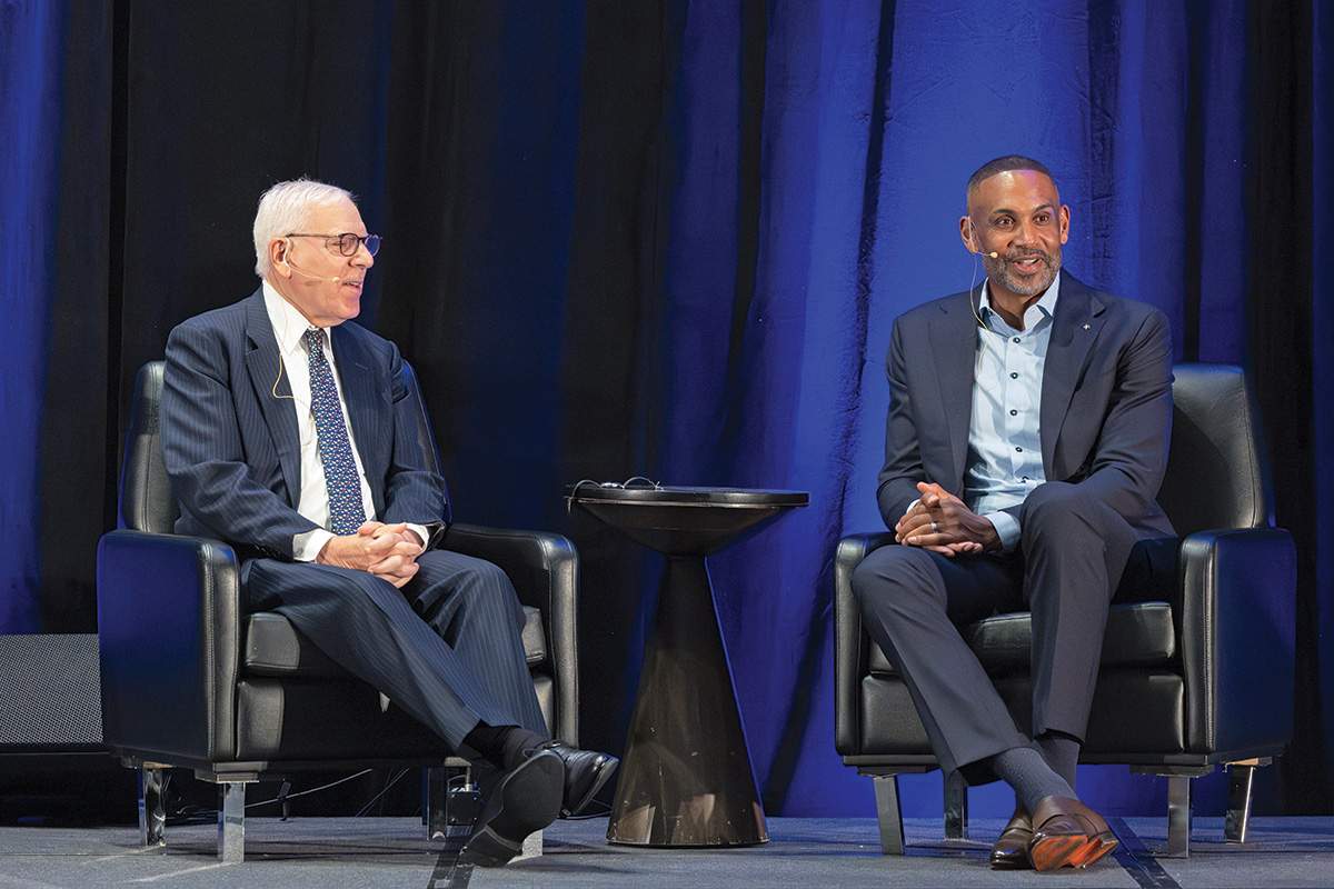 A photo of two people seated on a stage with a small round table between them.  On the left is David M. Rubenstein, a light-skinned man, wearing a dark suit and tie, who is looking at the other seated individual. On the right is Grant Hill, a dark-skinned man, wearing a business suit and looking out at the audience.