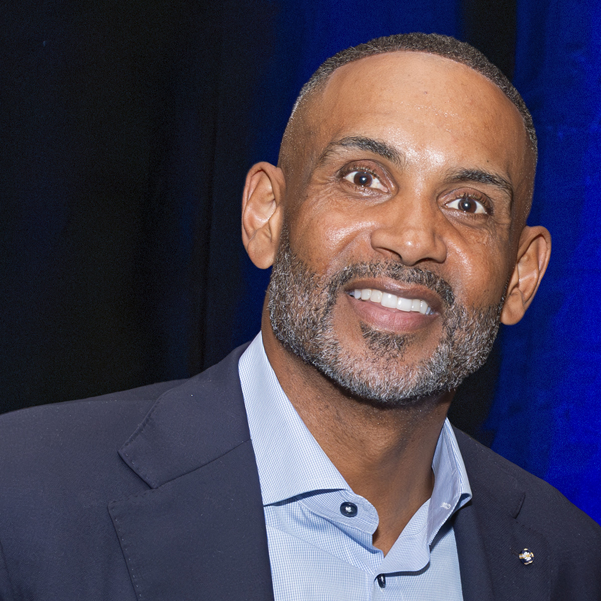 A photo of Grant Hill, a dark-skinned man with a beard and mustache, wearing a suit jacket and a blue business shirt, looking directly at the camera.