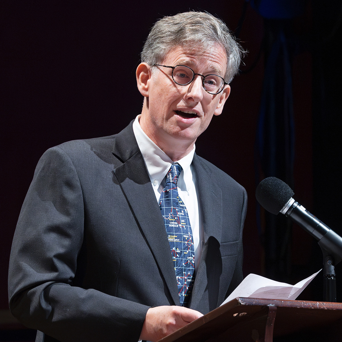 A photograph of Charles Gammie. He is wearing glasses and a business suit and tie. He is speaking to an audience, not shown.