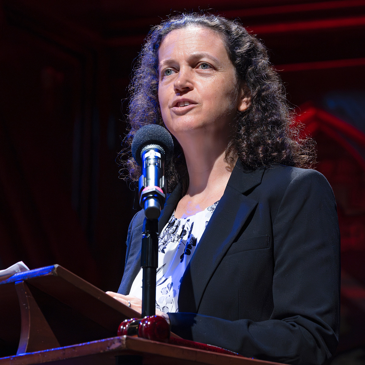 A photograph of Helene Muller-Landau, a woman with curly brown hair. She is speaking into a microphone and addressing an audience, not shown. 