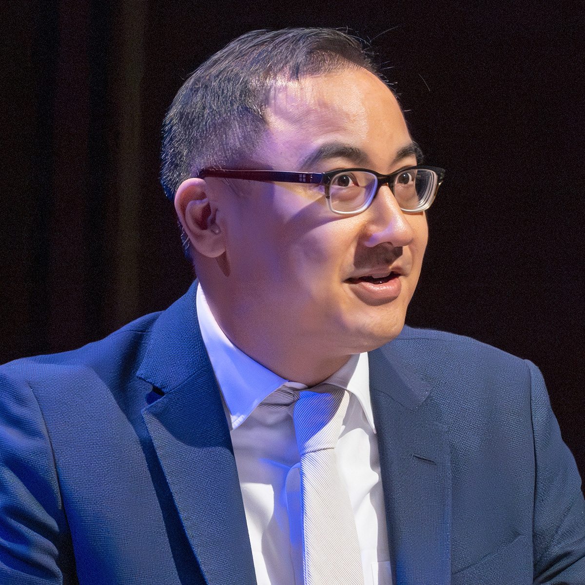 A photograph of Daniel Ho, a man with short black hair, wearing glasses, a suit, and tie. He is speaking at a podium and facing an audience not shown.  