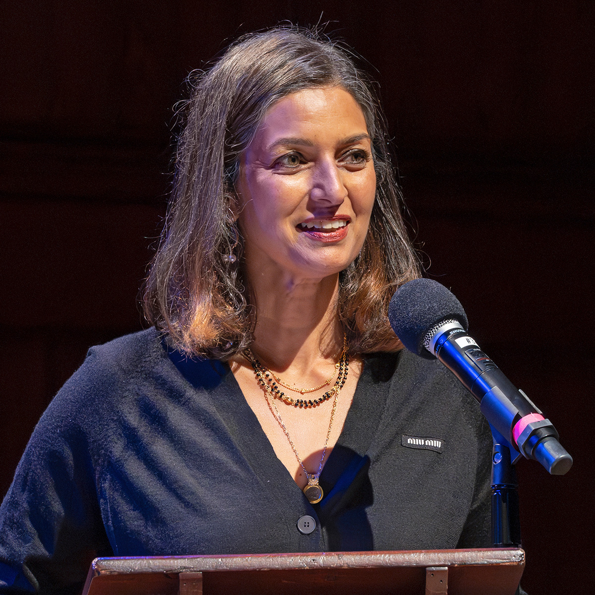 A photograph of Jhumpa Lahiri, a woman with shoulder-length brown hair. She is wearing a dark top and several gold necklaces. She is addressing an audience not shown. 