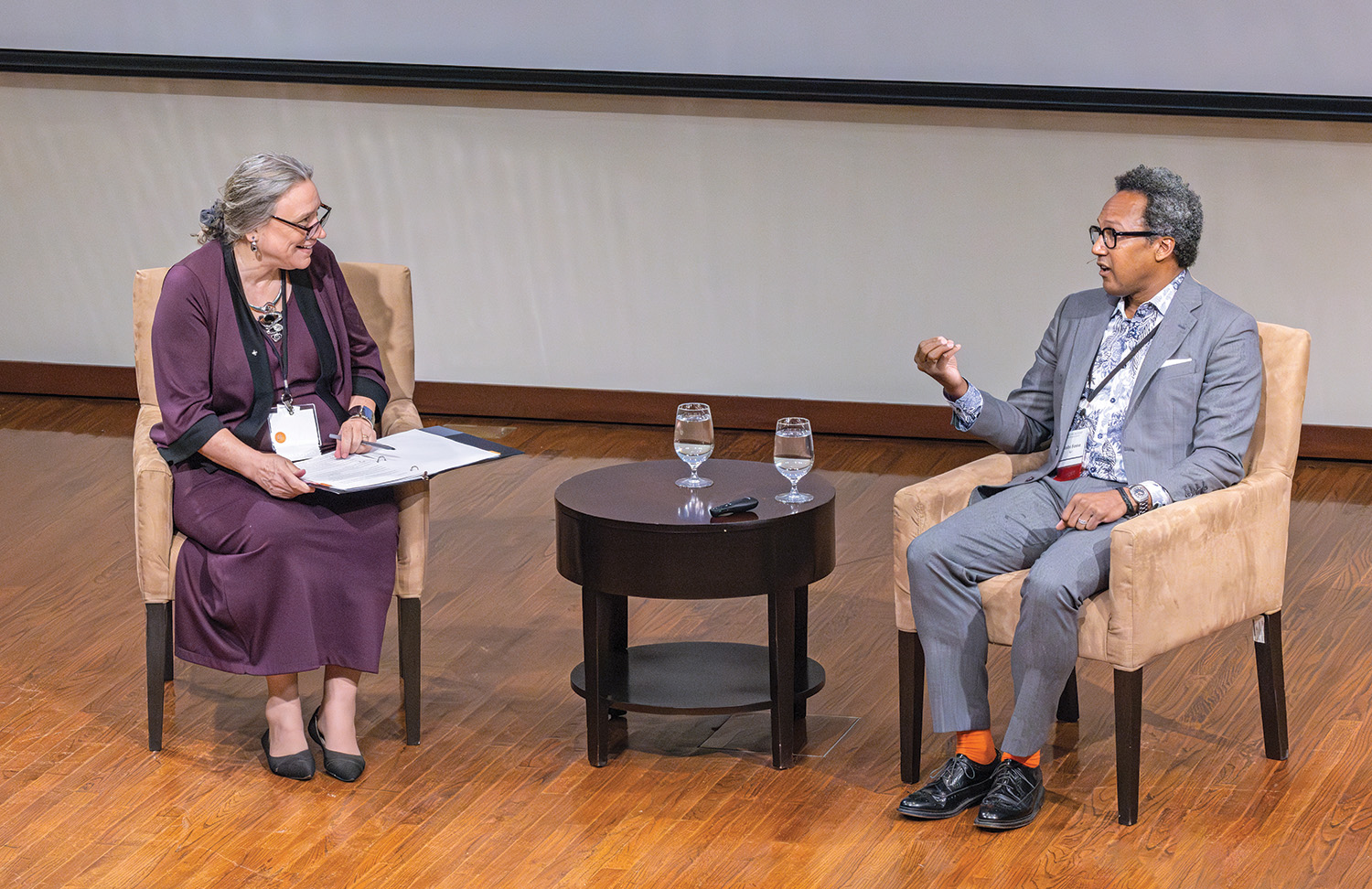 Two people in conversation with each other. The person on the left is seated in a chair and has a binder open on their lap. The person on the right is also seated in a chair and their right hand is raised as they make a point during the conversation.