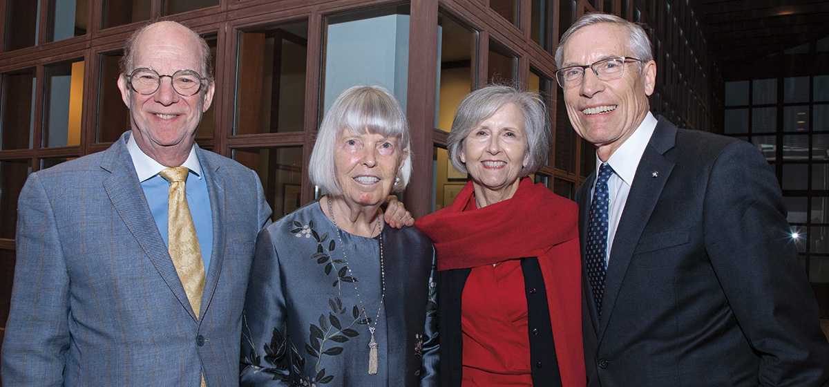 Four individuals stand together and smile at the viewer. On the far left is a man with fair skin, wearing glasses and a suit and tie. To his right is a woman with short gray hair, wearing a blue blouse. To her right is a woman with black and gray short hair, wearing a red scarf. At the far right is a man with fair skin and light gray hair, wearing glasses and a suit and tie.