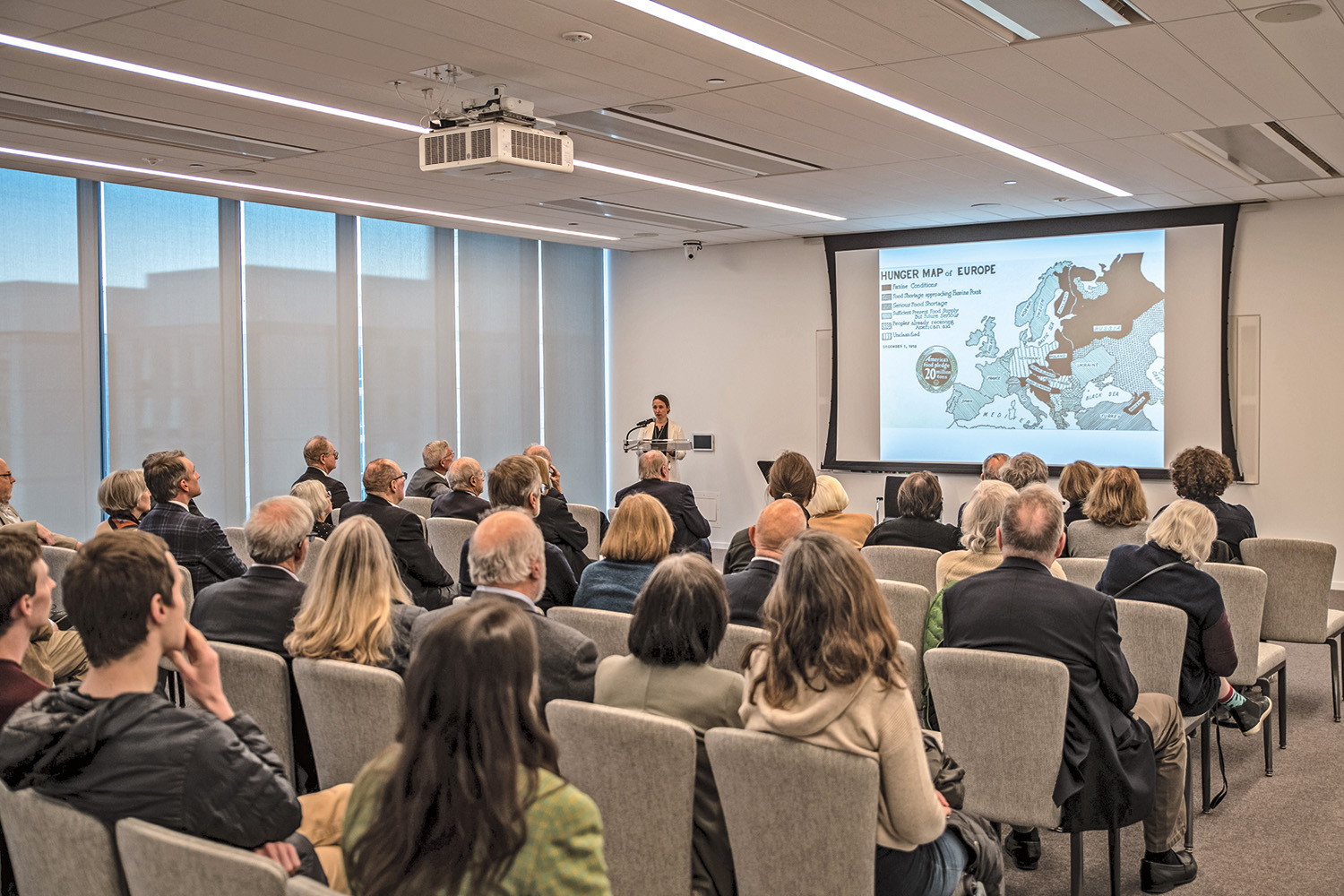 A full audience of seated people of all ages listen to a lecture from a speaker at a podium. The audience looks at a projected image of a hunger map of Europe. 