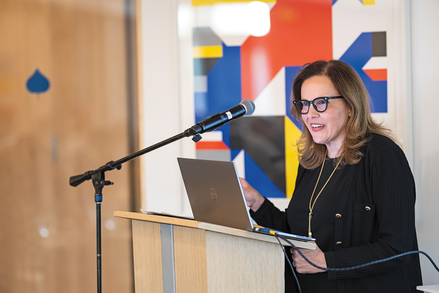 A woman in business attire is standing at a podium and speaking into a microphone. Behind her is a brightly patterned wall painting. 