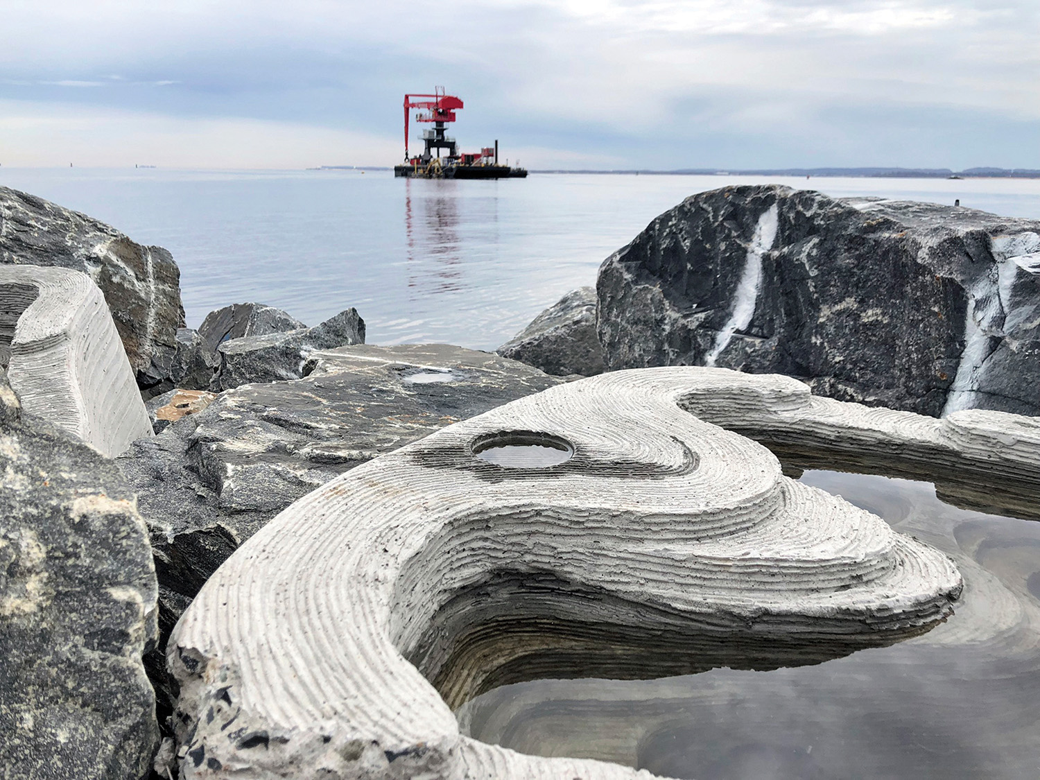 A manmade cement breakwater partially covered by water sits amid a rocky shore in the foreground. Red and gray machinery can be seen floating in the ocean in the background.  