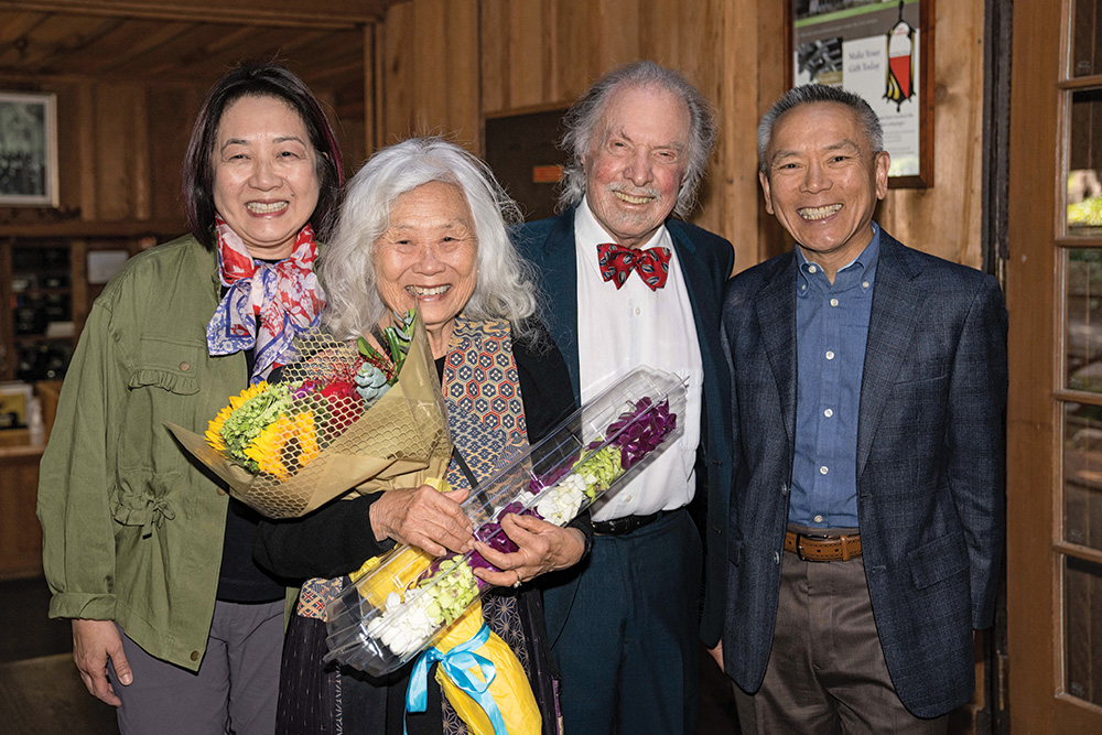 Maxine Hong Kingston stands beside three attendees at her award ceremony. Kingston holds two bouquets of flowers in her arms. All face the viewer and smile.