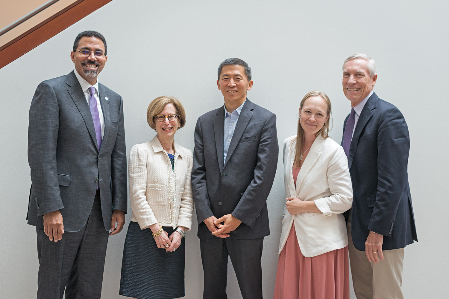 A group of five adults wearing business attire stand in front of a white wall and smile at the camera.