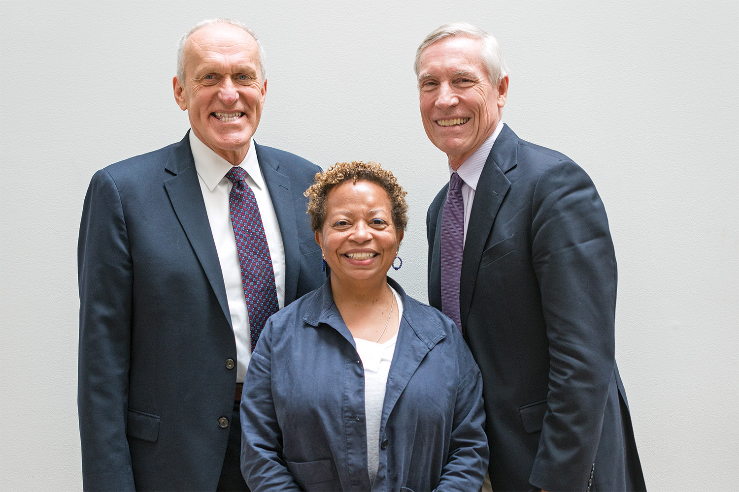 Three adults wearing business attire stand next to one another, in front of a white wall, and are smiling at the camera. 