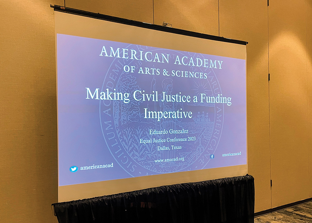 A screen shows a projection of a slide from a presentation. The slide reads: “American Academy of Arts and Sciences. Making Civil Justice a Funding Imperative.”