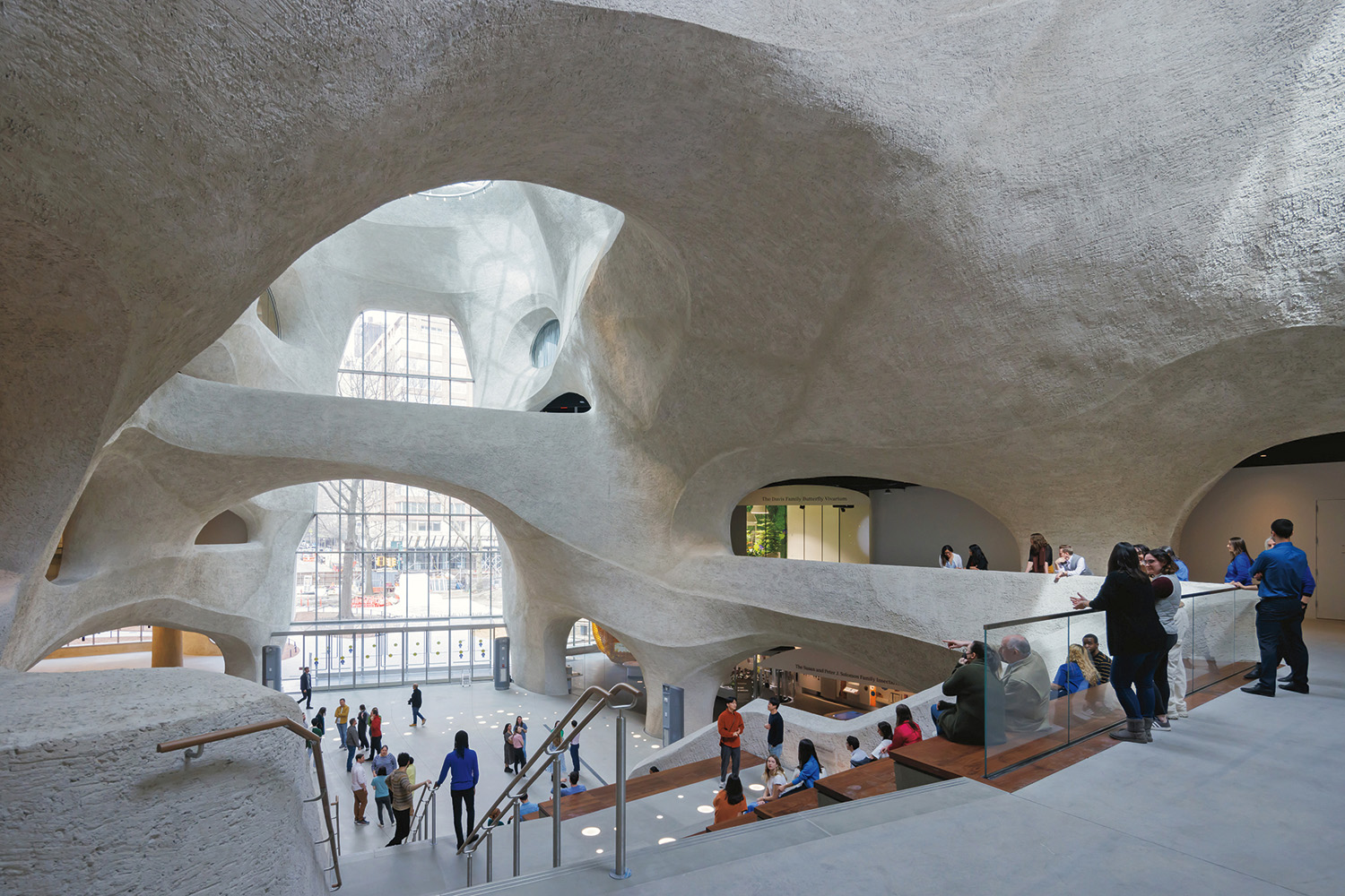 A photograph of the interior of a museum with cave-like architecture. People are exploring the space, which features flowing concrete walls and openings that resemble windows and portals. Natural light illuminates the space. 